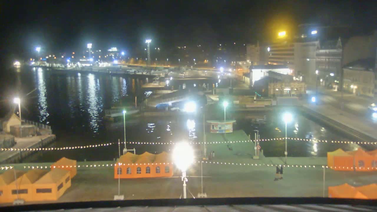 A nighttime view of a harbor with orange tents, lights illuminating the waterfront, and buildings in the background.