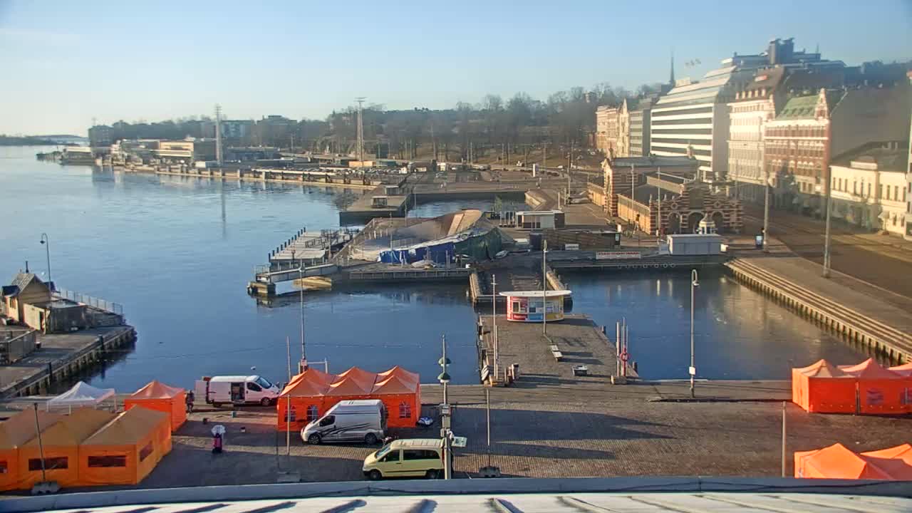 A harbor scene on a sunny day shows a calm body of water with several boats and docks, buildings in the background, and orange tents set up on a paved area.