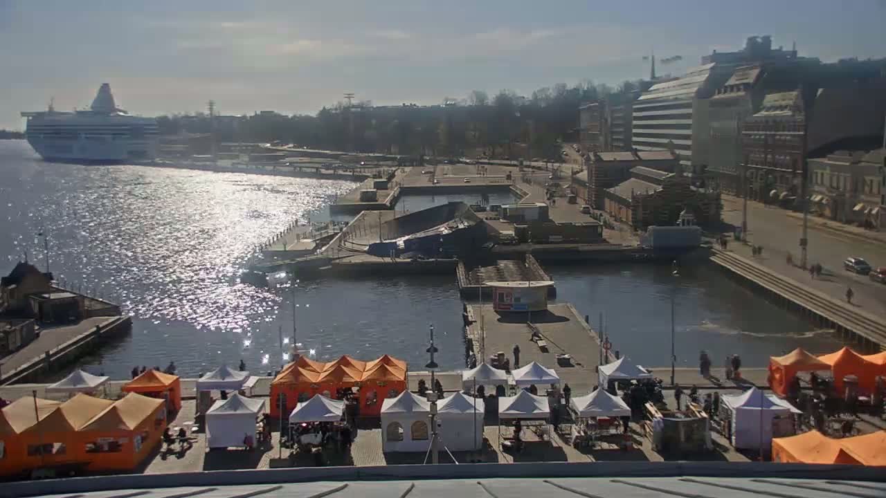 A sunny day overlooking a harbor with a large cruise ship in the distance, numerous market stalls set up along the waterfront, and several buildings lining the shore.