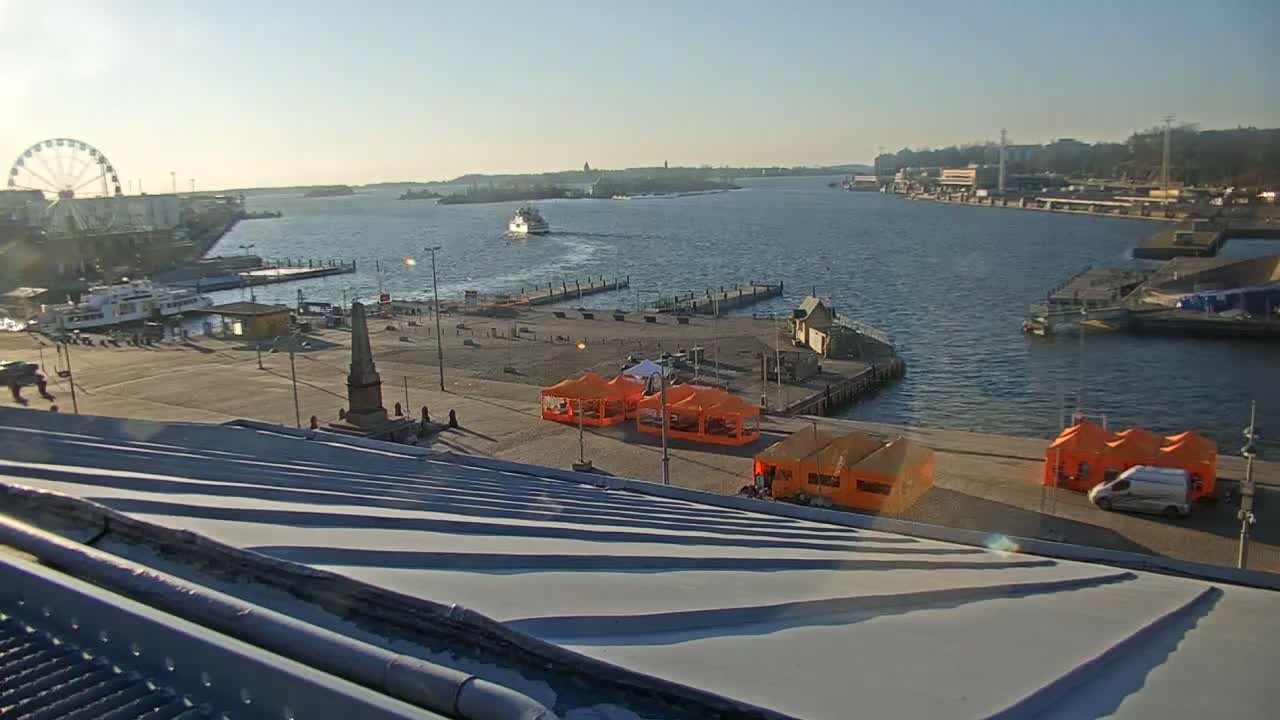 A sunny harbor scene shows a Ferris wheel, boats on the water, a monument, and several orange tents on a paved area next to a metal roof.