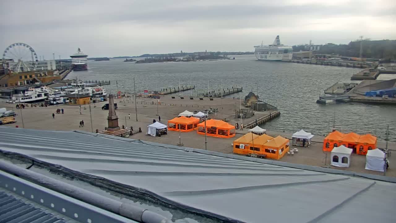 An overcast day reveals a harbor scene with a large ferry and several smaller boats docked, alongside a plaza featuring orange and white market tents and a Ferris wheel in the distance.