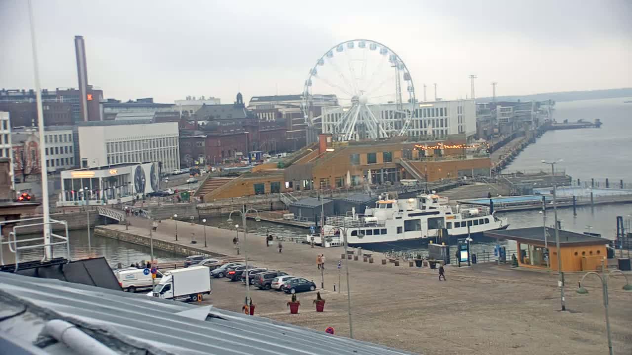 A harbor scene on an overcast day features a Ferris wheel, buildings, a ferry docked at a pier, and several cars parked nearby.