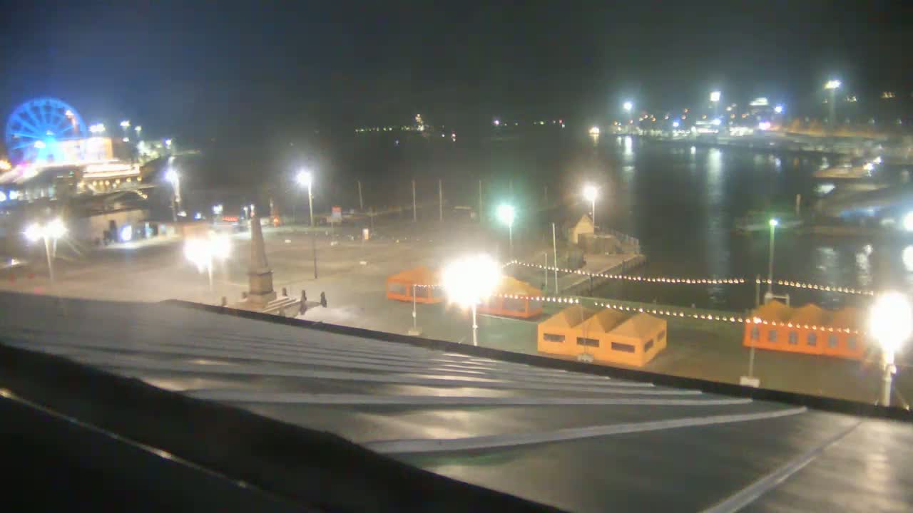 A nighttime view from a rooftop shows a harbor with boats, a Ferris wheel, a monument, and several small orange structures illuminated by bright lights.