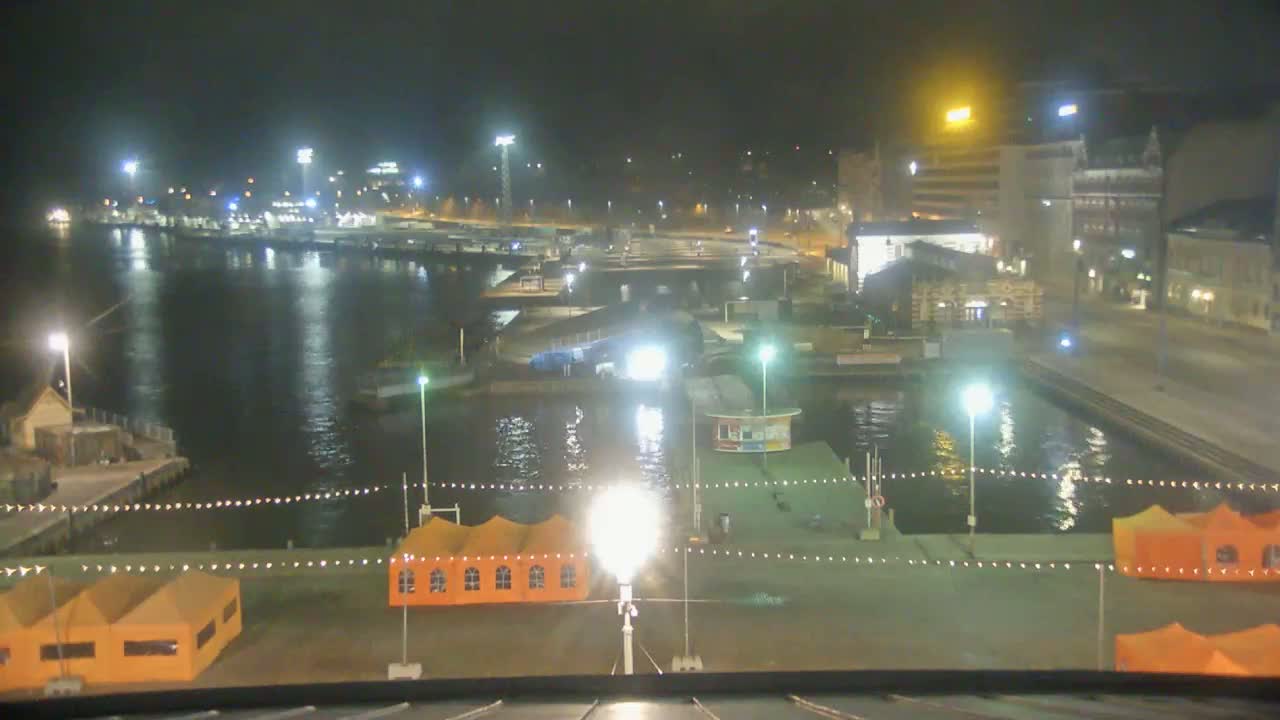 A nighttime view of a harbor with orange tents, lights illuminating the dock and surrounding buildings, and calm water reflecting the city lights.