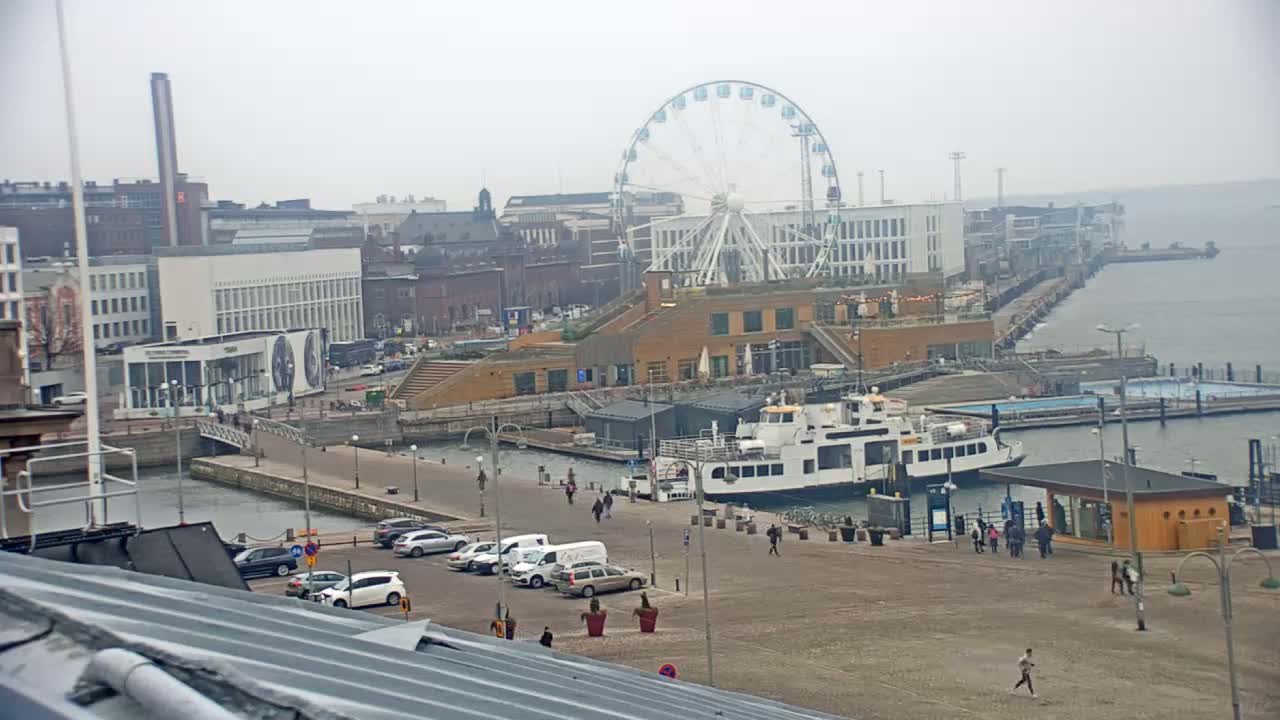 A waterfront cityscape on an overcast day features a Ferris wheel, buildings, a ferry, and parked cars, with a few pedestrians visible.