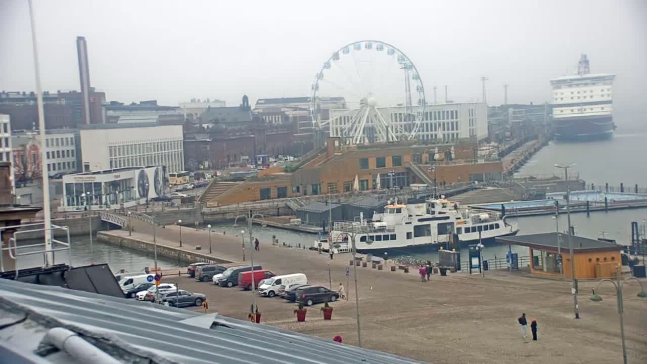 A ferry is docked near a Ferris wheel at a harbor in foggy conditions, with cars parked along a paved area.