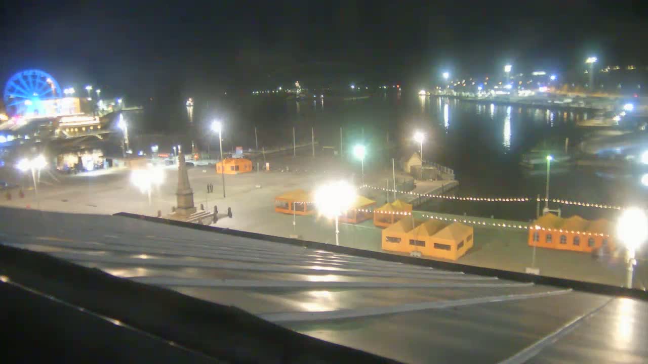 A nighttime view of a harbor at night, featuring a Ferris wheel, several orange tents, and a monument in a plaza, with calm water reflecting lights.