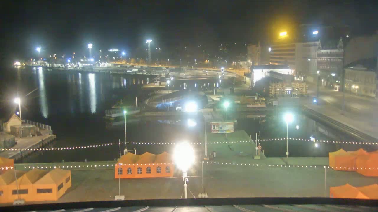 A nighttime view of a harbor with boats, buildings, and orange tents illuminated by numerous lights.