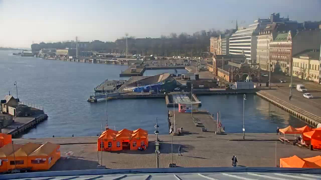 A harbor scene on a sunny day shows a body of water with boats, docked ships, buildings lining the waterfront, and several orange tents set up on a paved area.