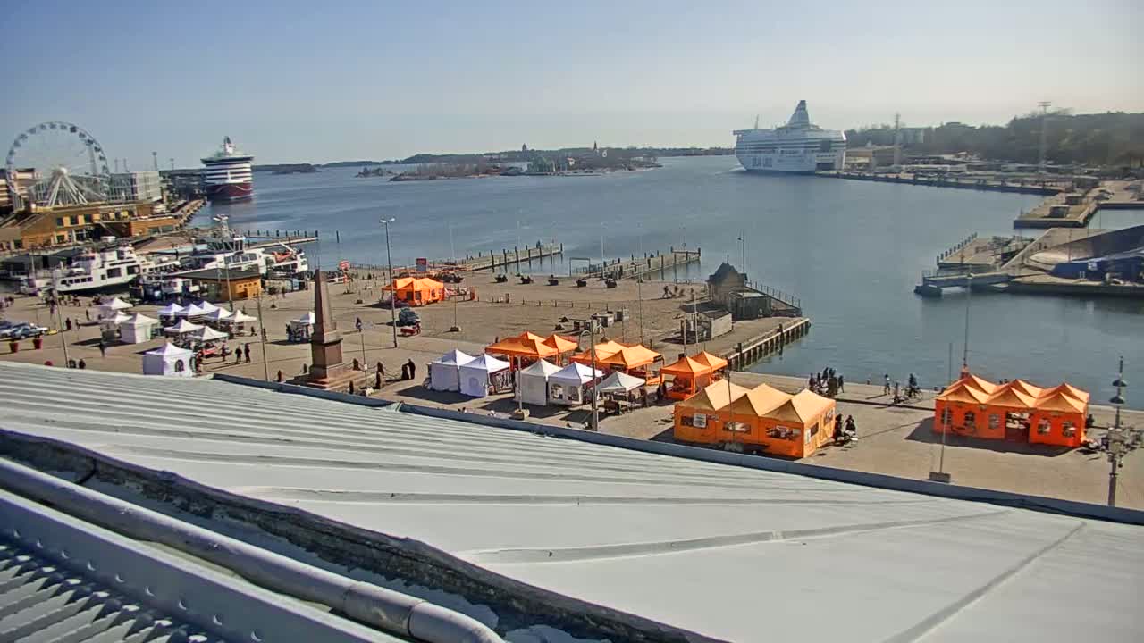 A harbor scene on a sunny day shows a Ferris wheel, several boats, a large cruise ship, and numerous orange and white market tents set up on a paved area near a waterfront.