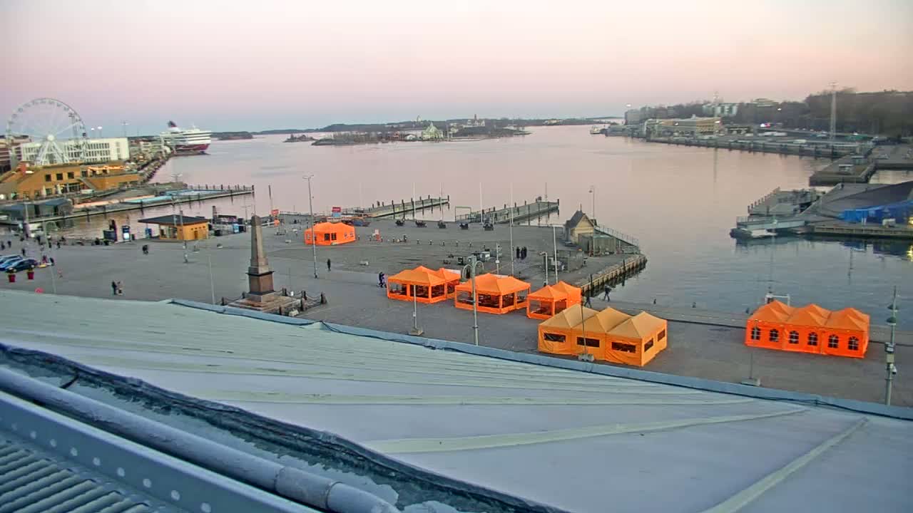 An elevated view of a harbor at dusk, showing several orange tents set up in a plaza near a calm body of water with boats and a Ferris wheel in the background.