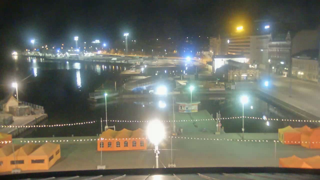 A nighttime view of a harbor with docked boats, orange tents, and brightly lit buildings across the water.