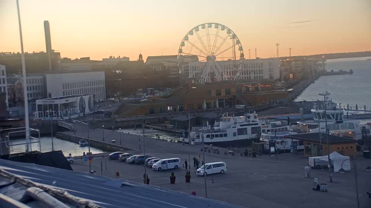 A waterfront city scene at sunset features a large Ferris wheel, docked boats, and several cars parked in a lot.