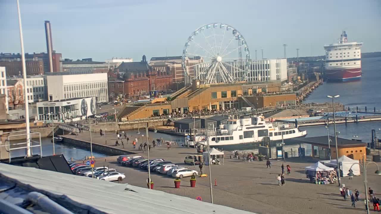 A sunny harbor scene features a Ferris wheel, a docked ferry, several buildings, and a large ship in the distance.