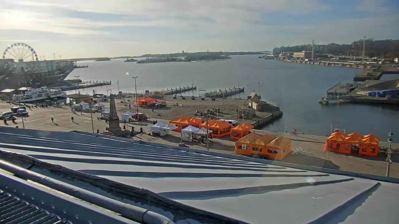 A harbor scene on a sunny day shows a Ferris wheel, boats, docked ships, several orange tents set up on a plaza, and a view across a body of water.
