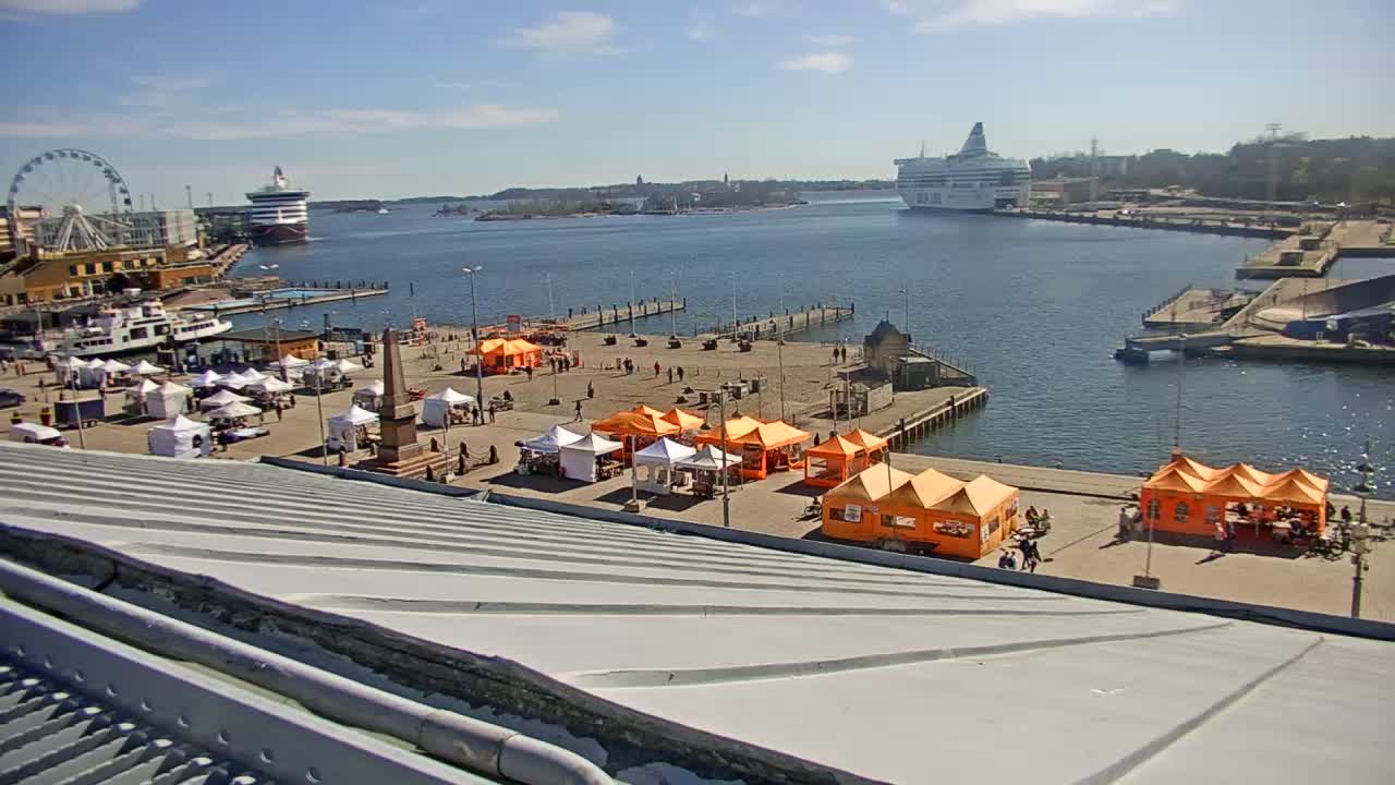 A harbor scene on a sunny day shows numerous market stalls, a Ferris wheel, and a large ferry docked in the distance, all viewed from a high vantage point.