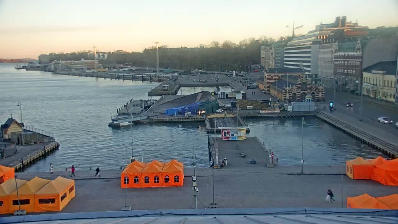 A harbor scene at dusk shows a calm body of water with piers, boats, buildings, and several orange tents on a paved area.