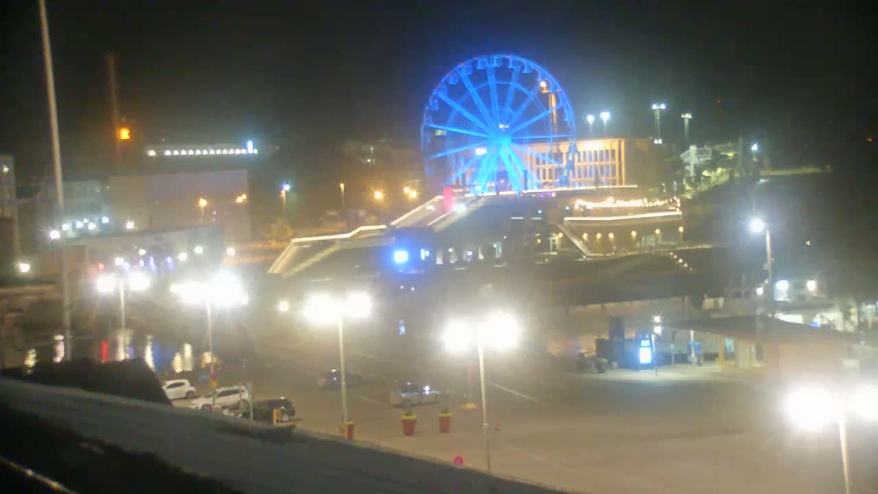 A brightly lit Ferris wheel stands out against a dark night sky above a parking lot with several cars and streetlights.