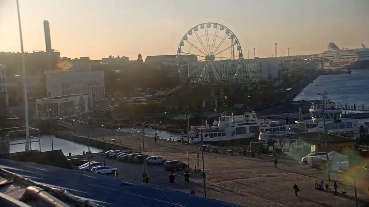 A waterfront scene at sunset shows a Ferris wheel, several docked ferries, cars parked along a quay, and buildings in the background.