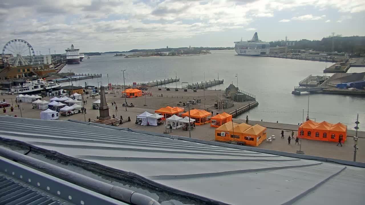 An overcast day shows a harbor with several ships, a Ferris wheel, and numerous market stalls set up on a plaza.