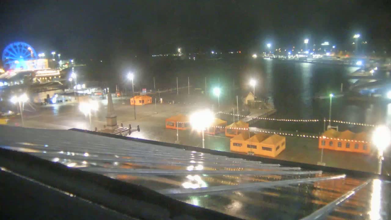 A rain-slicked rooftop overlooks a nighttime harbor scene with a Ferris wheel, illuminated tents, and a body of water under a dark sky.
