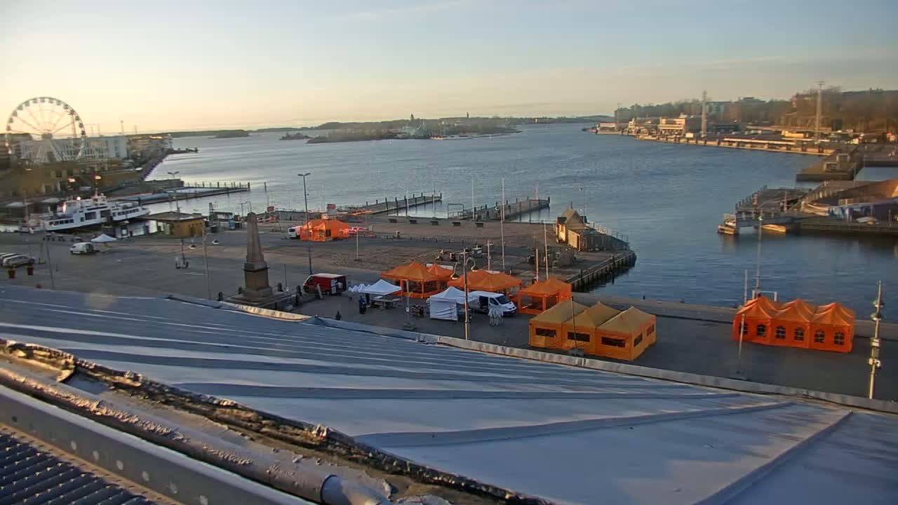 An elevated view of a harbor at sunrise shows a calm body of water with several boats, a Ferris wheel, and numerous orange tents set up on a paved area near a pier.