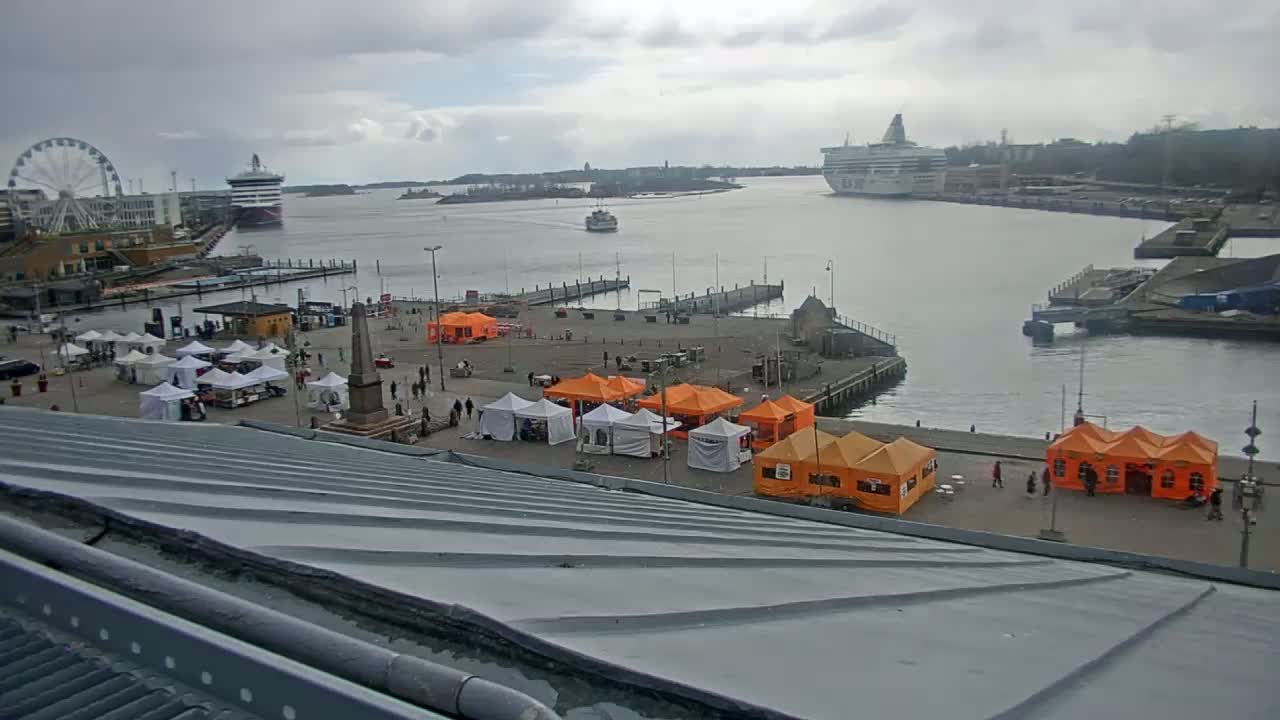 A harbor scene on an overcast day shows a Ferris wheel, several ships, a square with market stalls, and a metallic roof in the foreground.