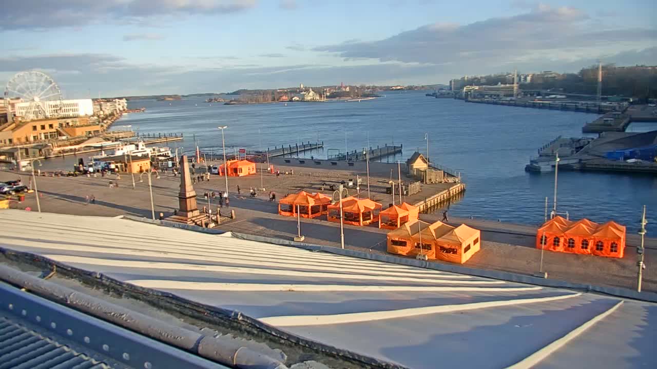 A harbor scene on a partly cloudy day shows a waterfront area with several orange tents, a Ferris wheel in the distance, and boats moored at the docks.
