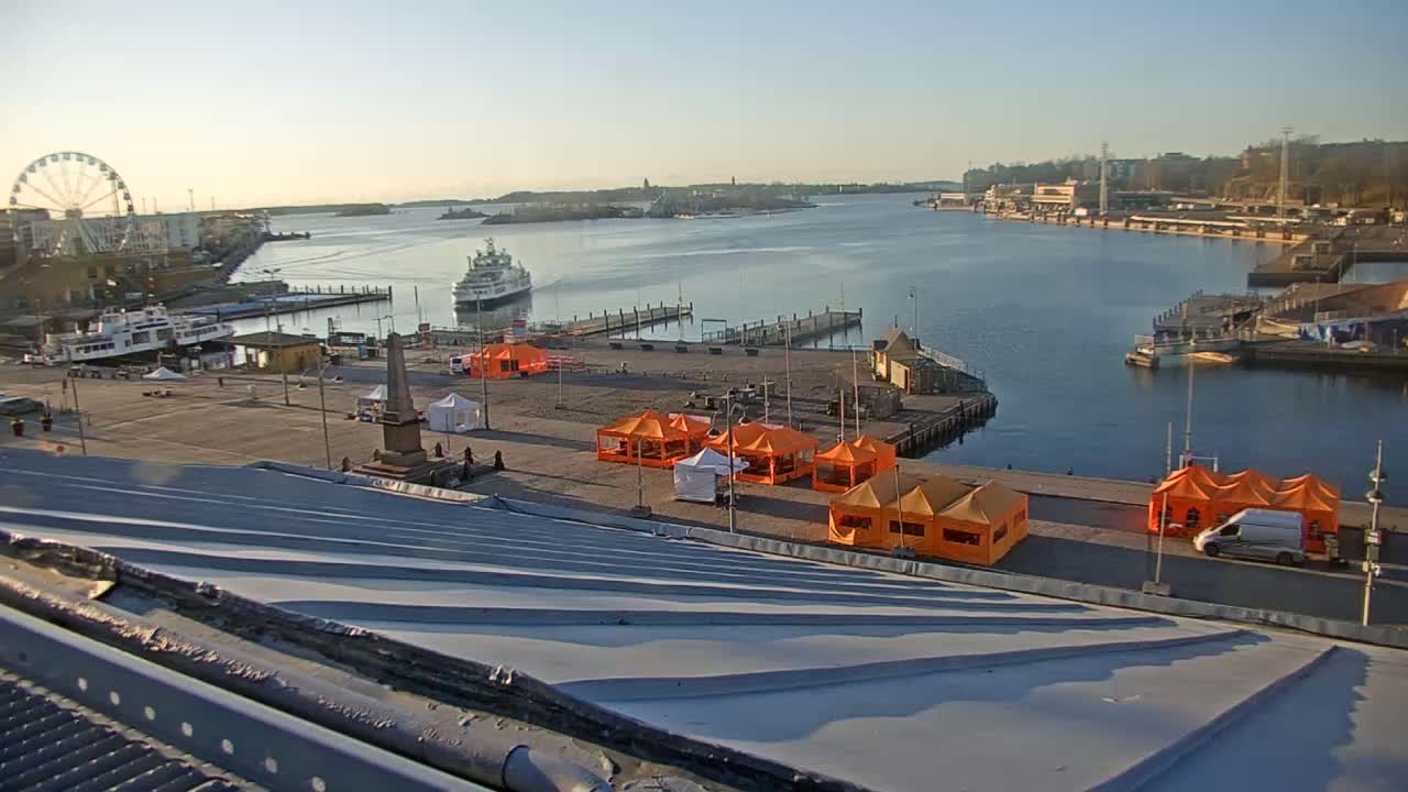 A harbor scene on a sunny day shows a ferry, several smaller boats, a Ferris wheel, and numerous orange tents set up on a paved area near a waterfront, viewed from a sloped metal roof.