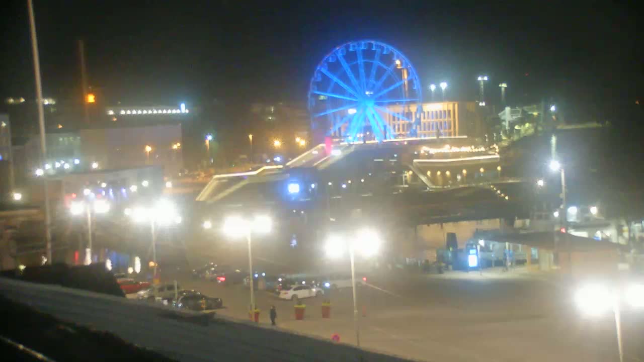A brightly lit Ferris wheel, illuminated in blue, stands prominently at night in a waterfront area with parked cars and streetlights.