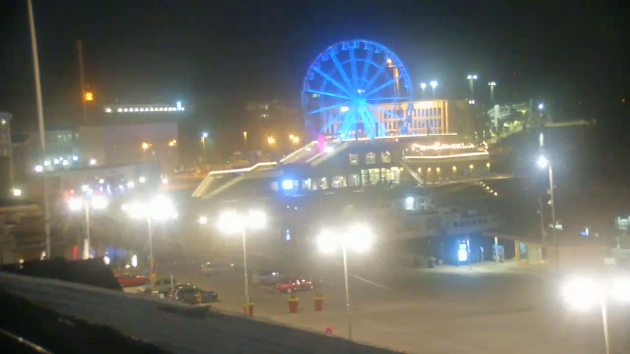 A brightly lit Ferris wheel dominates a nighttime scene of a waterfront area with buildings, streetlights, and a few cars.