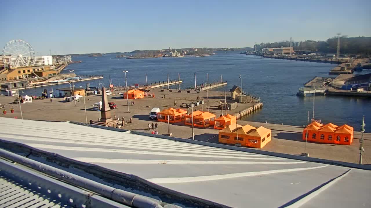 A harbor scene on a sunny day shows several orange tents set up on a paved area near a body of water with docked piers, a Ferris wheel in the background, and a view of a city across the water.