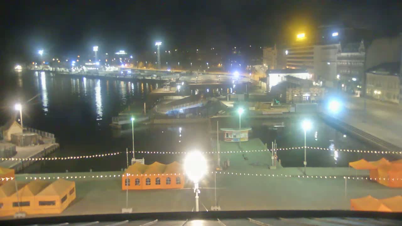 A nighttime view of a harbor with boats moored, illuminated docks, and several orange tents on the waterfront.