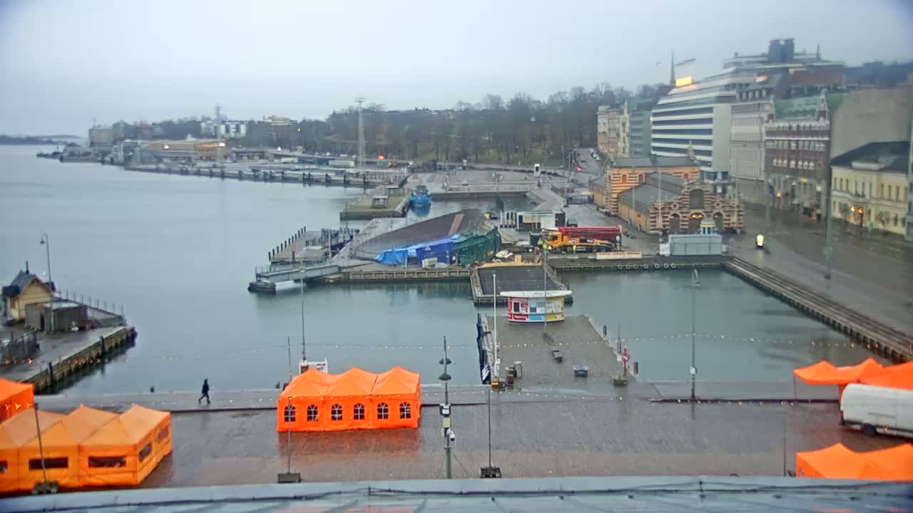 A harbor scene on an overcast day shows several boats, buildings, and orange tents set up on a wet dock.