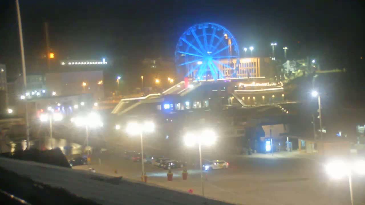 A brightly lit Ferris wheel, illuminated in blue, stands prominently at night over a paved area with cars and streetlights.