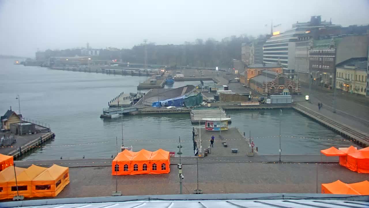 A harbor scene on an overcast day shows a partly covered structure on a dock, several orange tents on a plaza, and buildings in the background.