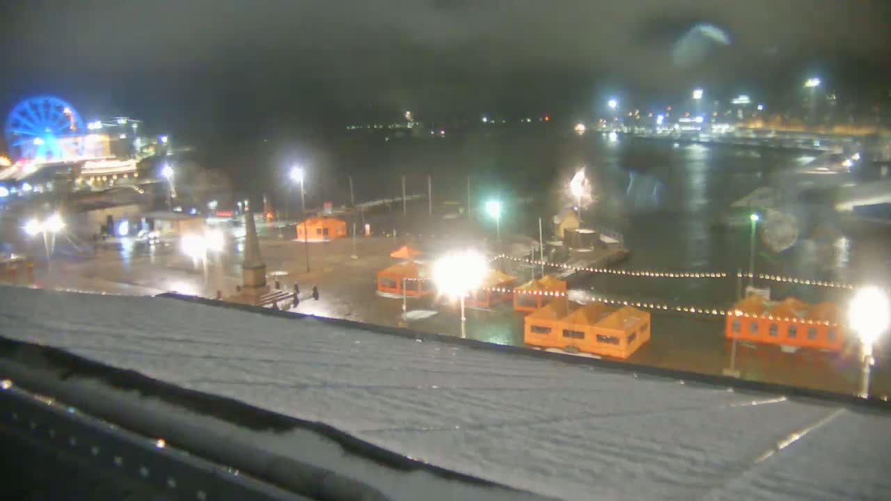 A nighttime view of a snow-covered waterfront area with illuminated orange structures, a Ferris wheel, and a body of water, under overcast conditions.