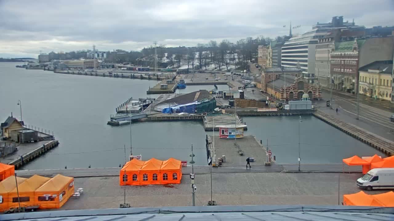 A harbor scene on an overcast day shows a city waterfront with docked boats, buildings, and several orange tents on a paved area.