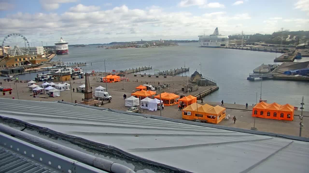 A harbor scene on a partly cloudy day shows a Ferris wheel, several ships, numerous white and orange tents set up on a plaza, and a gray metal roof in the foreground.