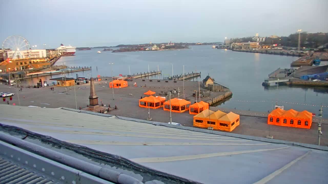 An elevated view of a harbor scene on a partly cloudy day, showing a waterfront square with several orange tents, a Ferris wheel, ships, and buildings.