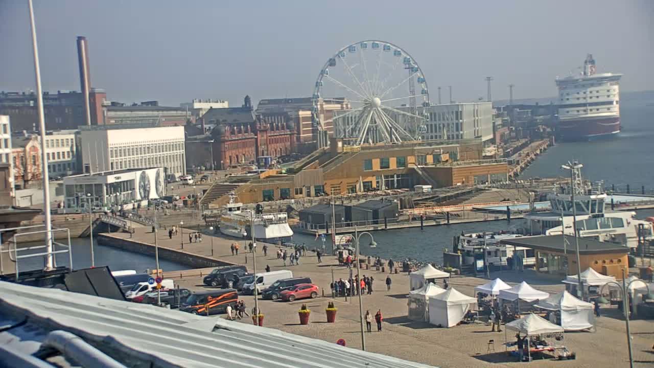 A harbor scene on a sunny day shows a Ferris wheel, buildings, boats, and several market tents with people walking around.
