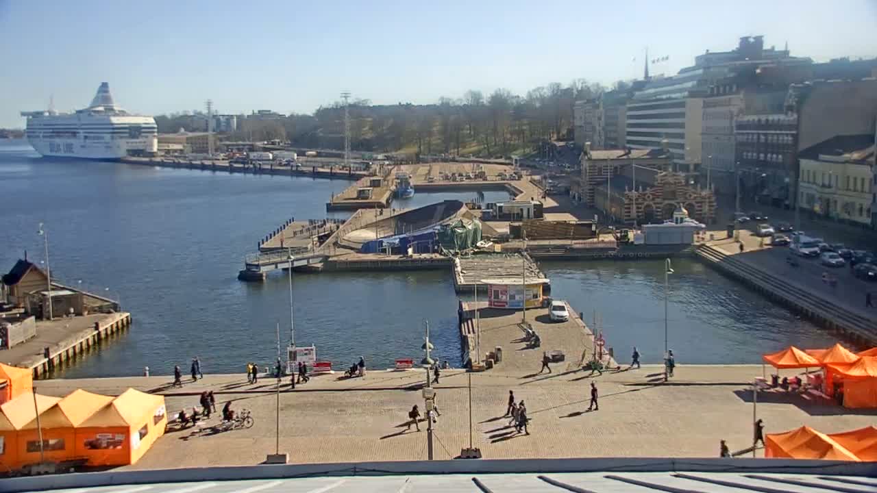 A harbor scene on a sunny day shows a ferry docked, construction work underway at a pier, people walking on paved areas near several orange tents, and buildings lining the waterfront.