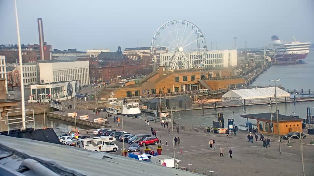 A harbor scene on an overcast day shows a Ferris wheel, buildings, docked boats, cars in a parking lot, and people walking along a paved area.