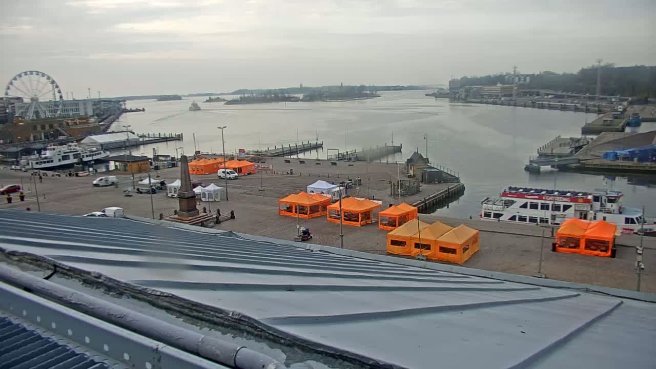 An overcast day shows a harbor scene with several boats, a Ferris wheel in the distance, and numerous orange tents set up on a paved area near the water.
