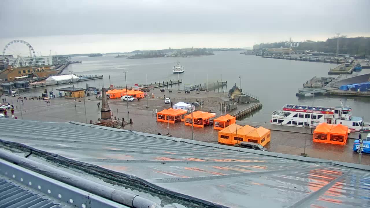 A harbor scene on an overcast day shows several orange market stalls set up on a wet paved area near a dock with boats, a Ferris wheel in the background.