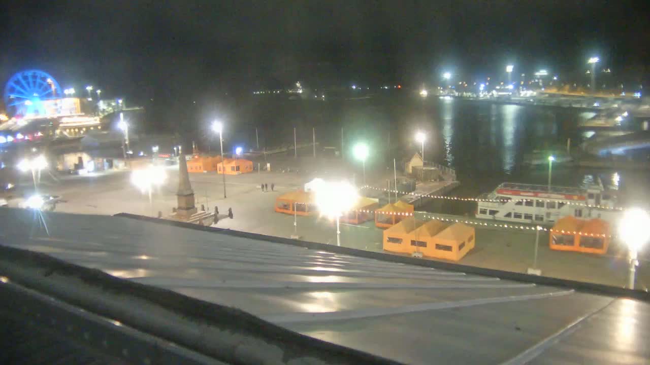 A nighttime view of a harbor area with illuminated tents, a Ferris wheel, and a docked boat, under clear skies.