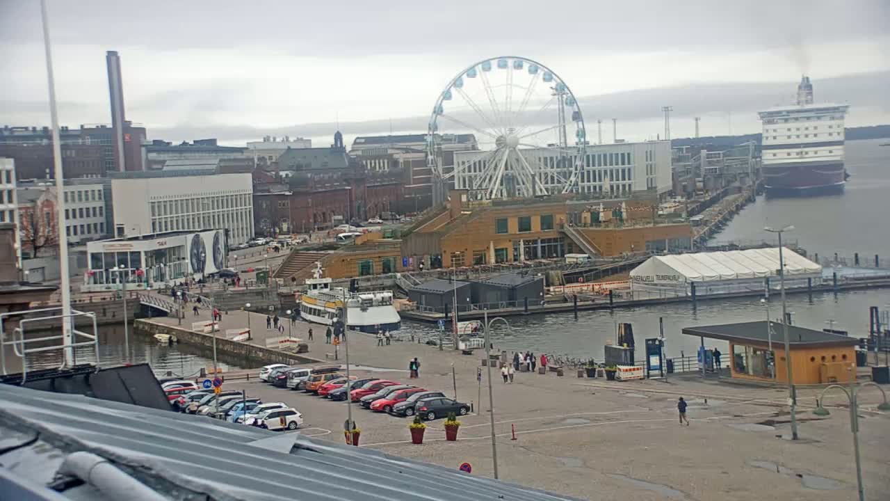 A city harbor scene on an overcast day shows a Ferris wheel, several buildings, a large cruise ship, smaller boats, cars in a parking lot, and a few pedestrians.