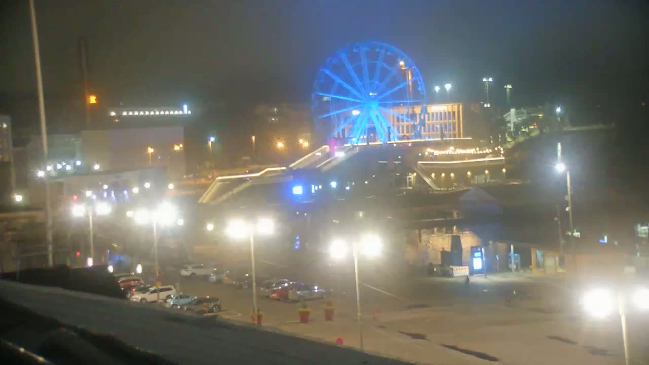 A brightly lit Ferris wheel glows blue under a hazy, night sky, overlooking a city street with parked cars and streetlights.