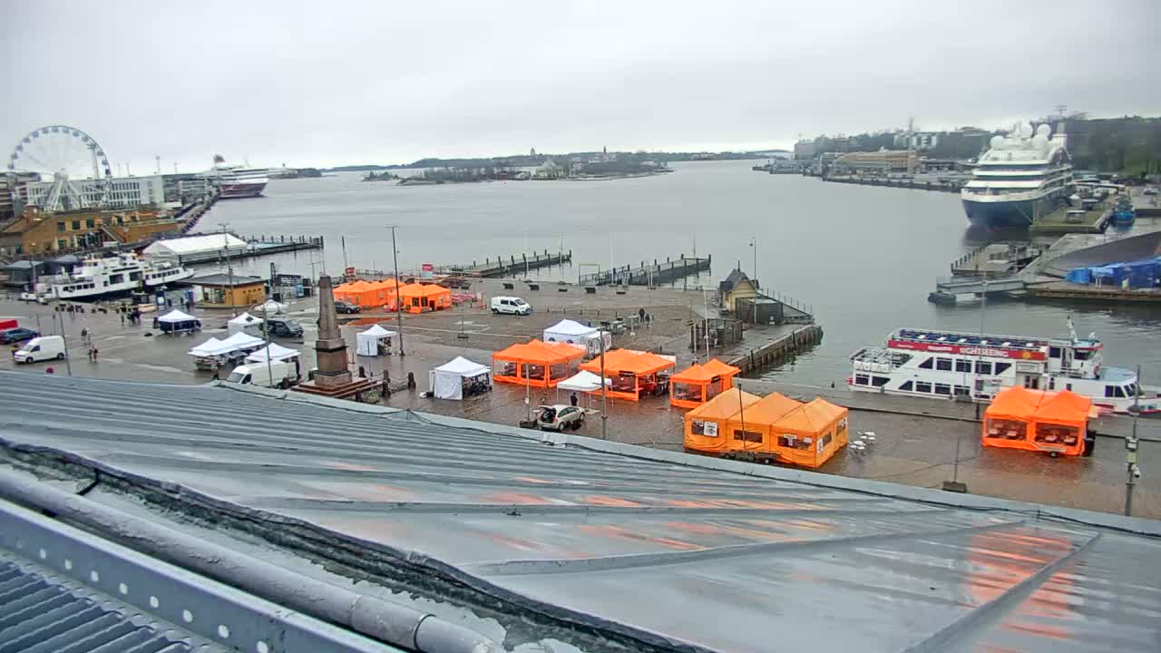 A harbor scene on an overcast day shows numerous orange and white tents set up on a waterfront plaza near docked boats, ferries, and a cruise ship, with a Ferris wheel visible in the background.