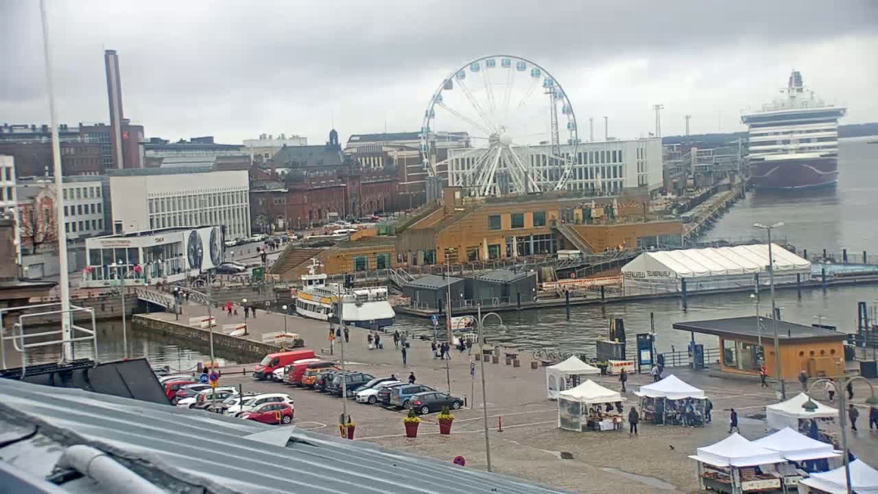 A waterfront city scene on an overcast day shows a Ferris wheel, a cruise ship, several buildings, a parking lot, market stalls, and boats in the harbor.